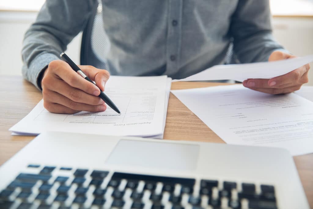 Personne annotant des documents sur un bureau, à côté d’un ordinateur portable